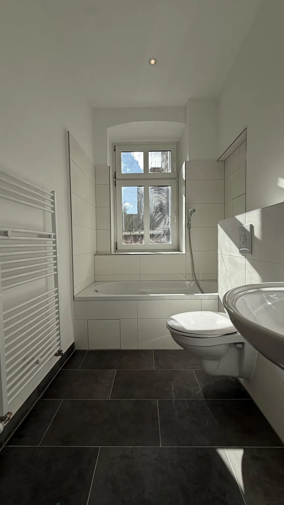 Fertiggestelltes Badezimmer mit Badewanne, WC, Waschbecken, weißen Wandfliesen und dunklem Boden